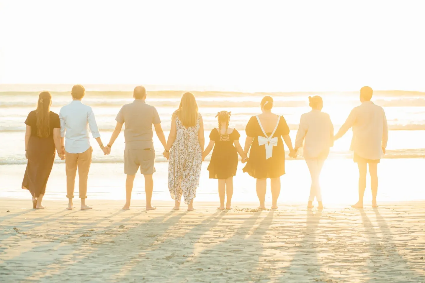 Backlit family of eight standing hand in hand at the water's edge, facing the low sun.