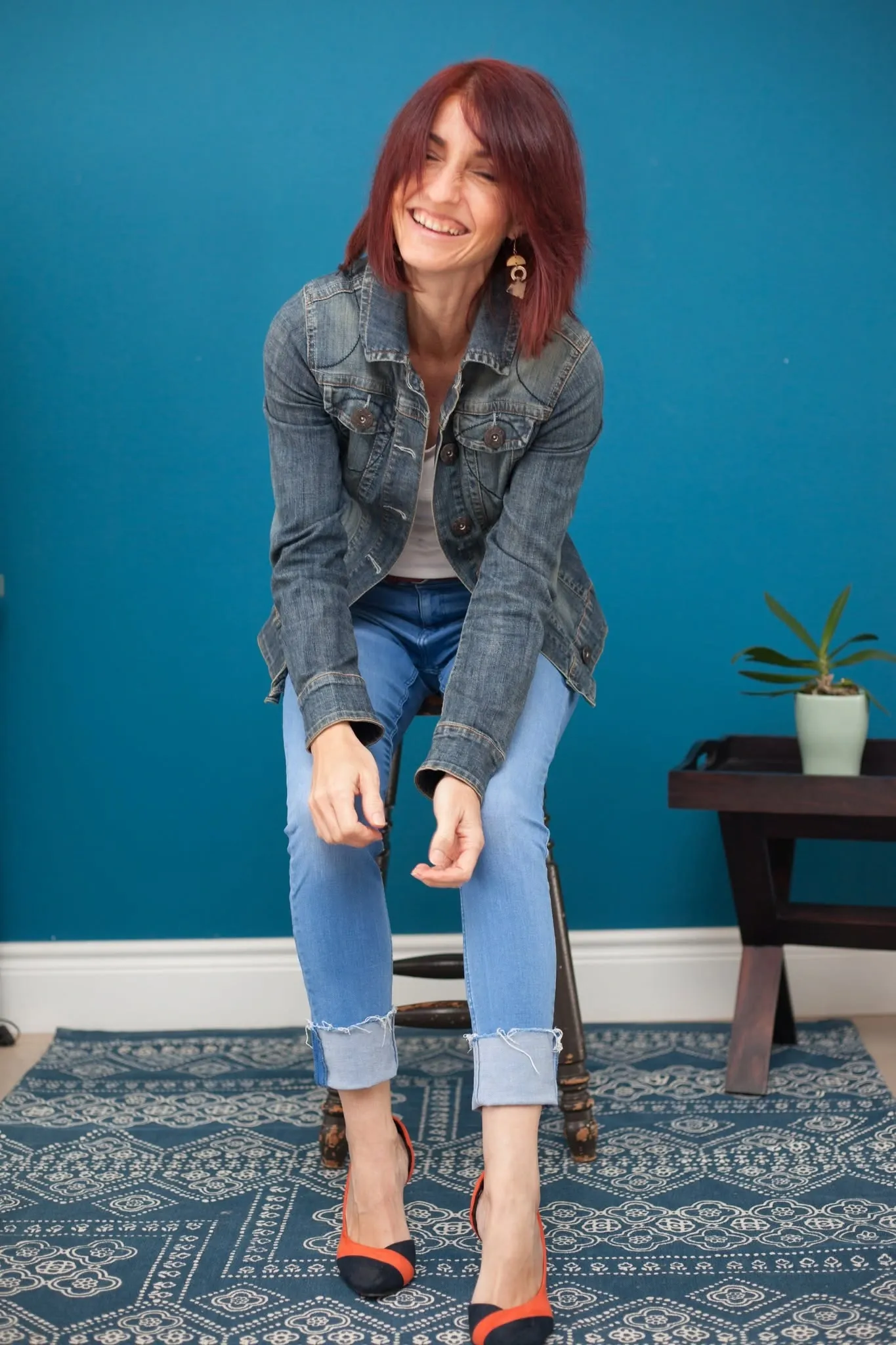 Full-length personal branding portrait of a laughing woman seated on a stool in front of a teal wall.