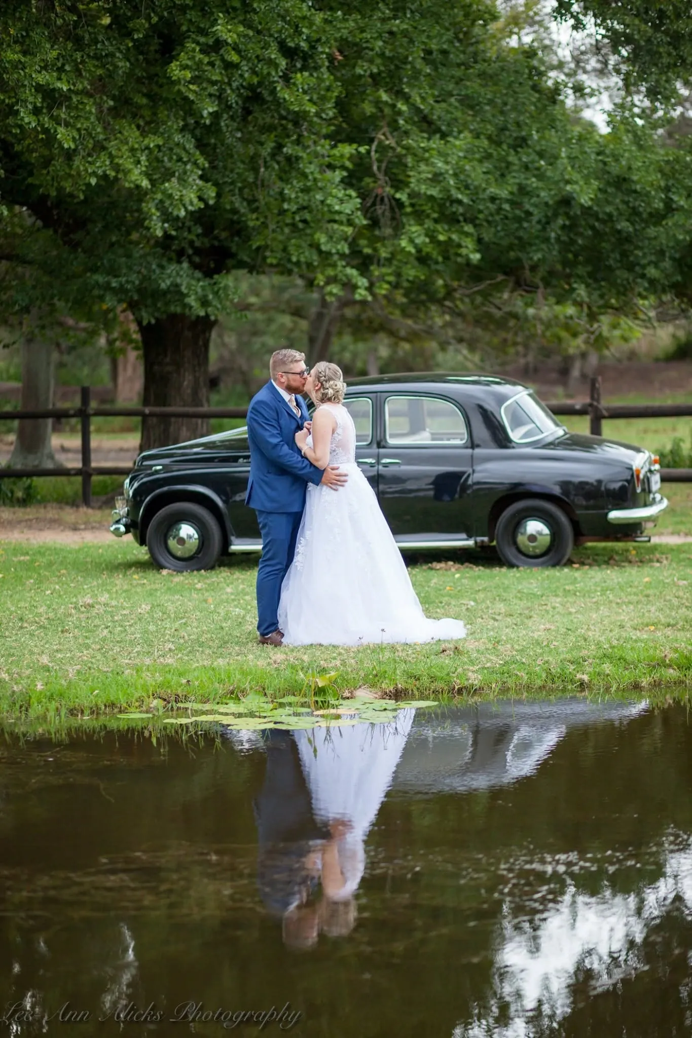 Bride and groom kissing beside a black vintage car, the whole scene reflected in a pond.